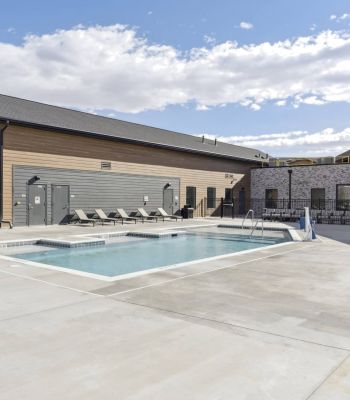 Outdoor swimming pool with lounge chairs and modern buildings under a partly cloudy sky.
